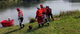 Na zdjęciu widoczni ratownicy stojący na brzegu akwenu Korzeń, zabezpieczeni w kamizelki ratunkowe oraz kask, przygotowujący się do rozpoczęcia ćwiczeń z zakresu ratownictwa wodnego. W tle stojąca zwodowana łódź.