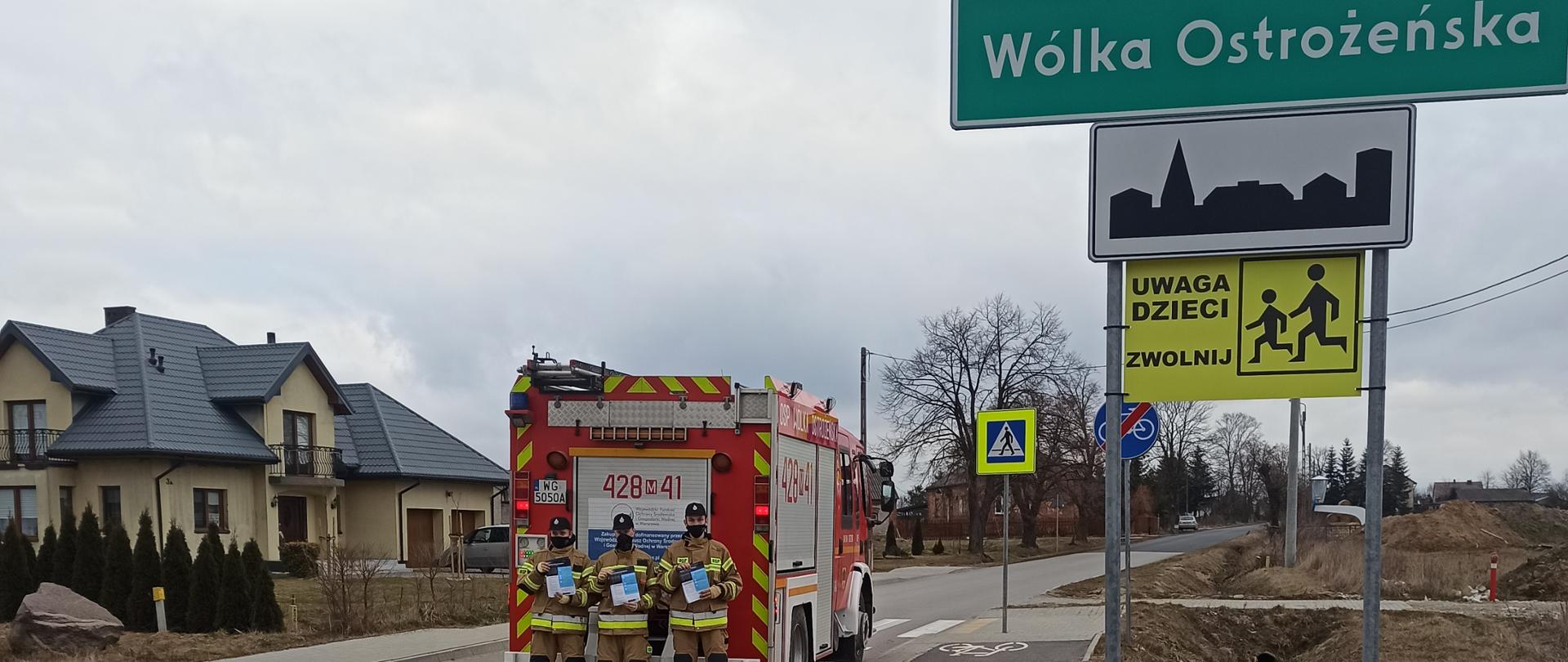 trzech strażaków stoi przed wozem strażackim na ulicy . Obok znajduje się znak z napisem Wólka Ostrożeńska a z tyłu samochodu zabudowania.