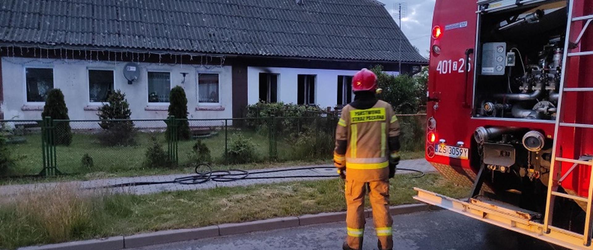 Zdjęcie przedstawia parterowy dom dwurodzinny w którym wybuch pożar. Przed płotem ogradzającym posesję stoi strażak w umundurowaniu specjalnym koloru piaskowego i czerwonym hełmem. Po prawej stronie samochód pożarniczy