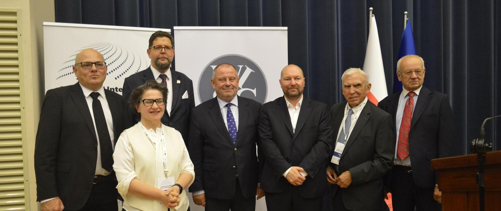 Przed przykrytą czarnym materiałem ścianą stoi grupa siedmiu osób, pośrodku z tyłu wiceminister Szeptycki.