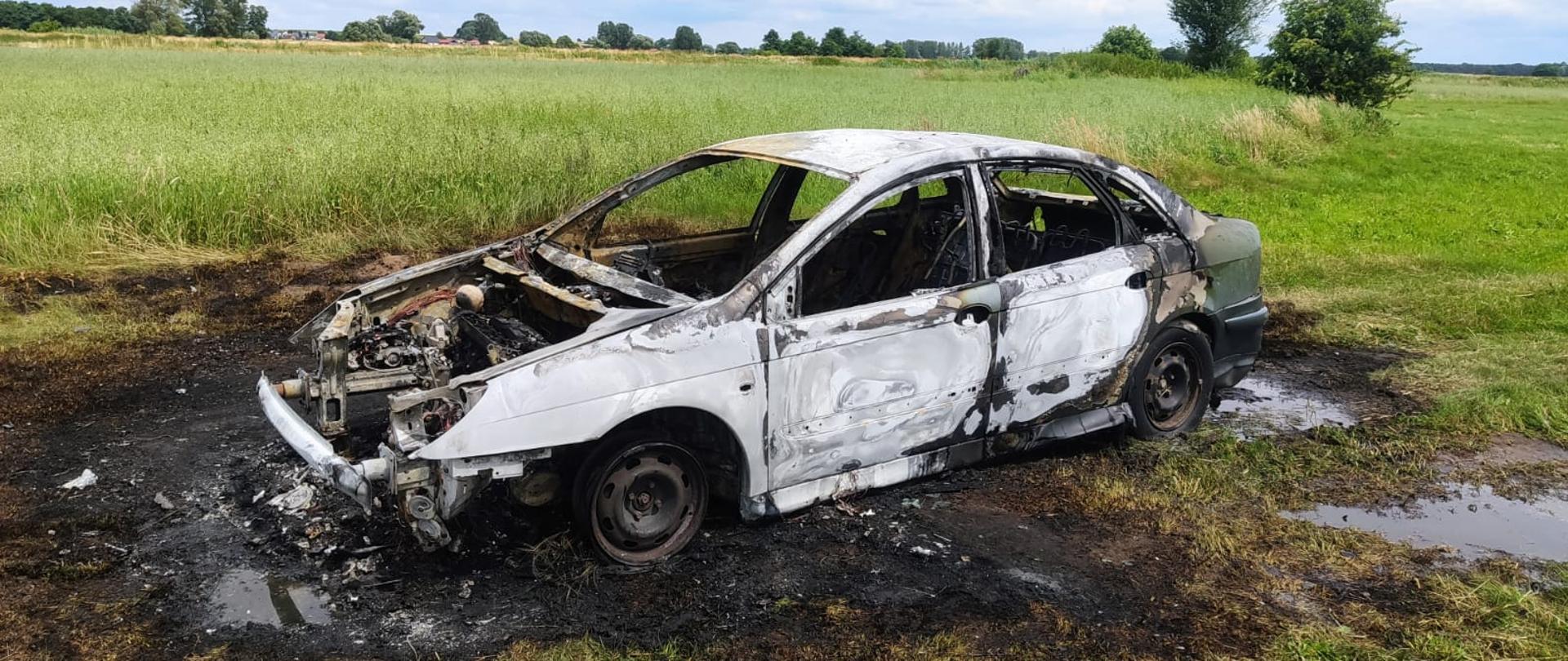 Zdjęcie przedstawia doszczętnie spalony samochód osobowy. Trawa otaczająca samochód jest również wypalona.