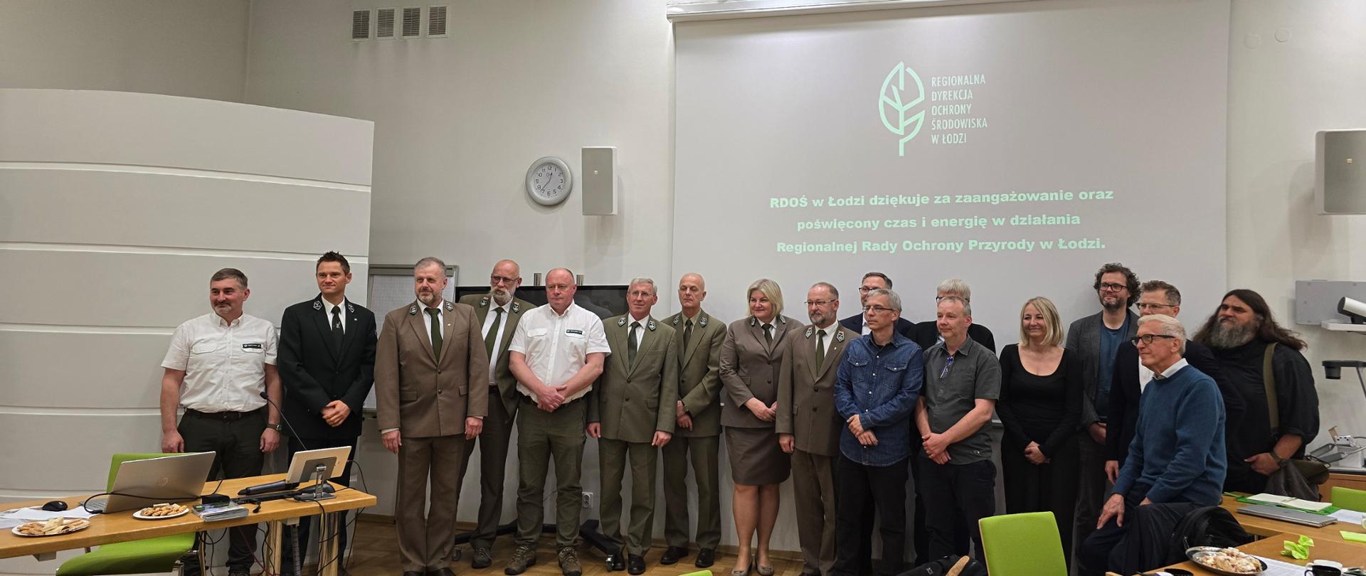 Grupa ludzi, część w ciemnozielonych mundurach w sali. Usmiechnięte twarze. Za nimi na rzutniku widoczne logo liśc i napis Regionalna Dyrekcja Ochrony Środowiska w Łodzi. Poniżej napis : RDOŚ w Łodzi dziekuje za zaangazowanie oraz poswiecony czas i energię w dzialania regionalnej Rady Ochrony Przyrody w Łodzi. 