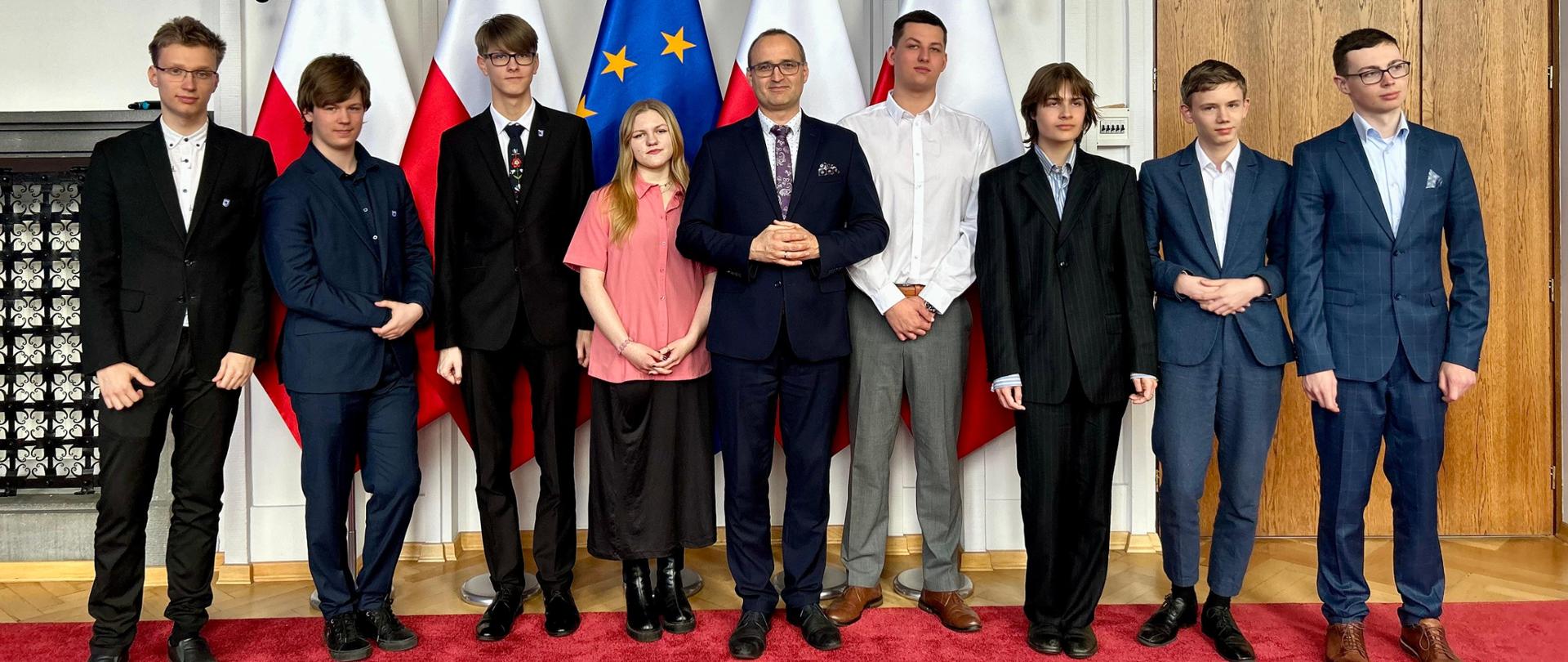 Grupa osób stoi na tle flag polskich i unijnej