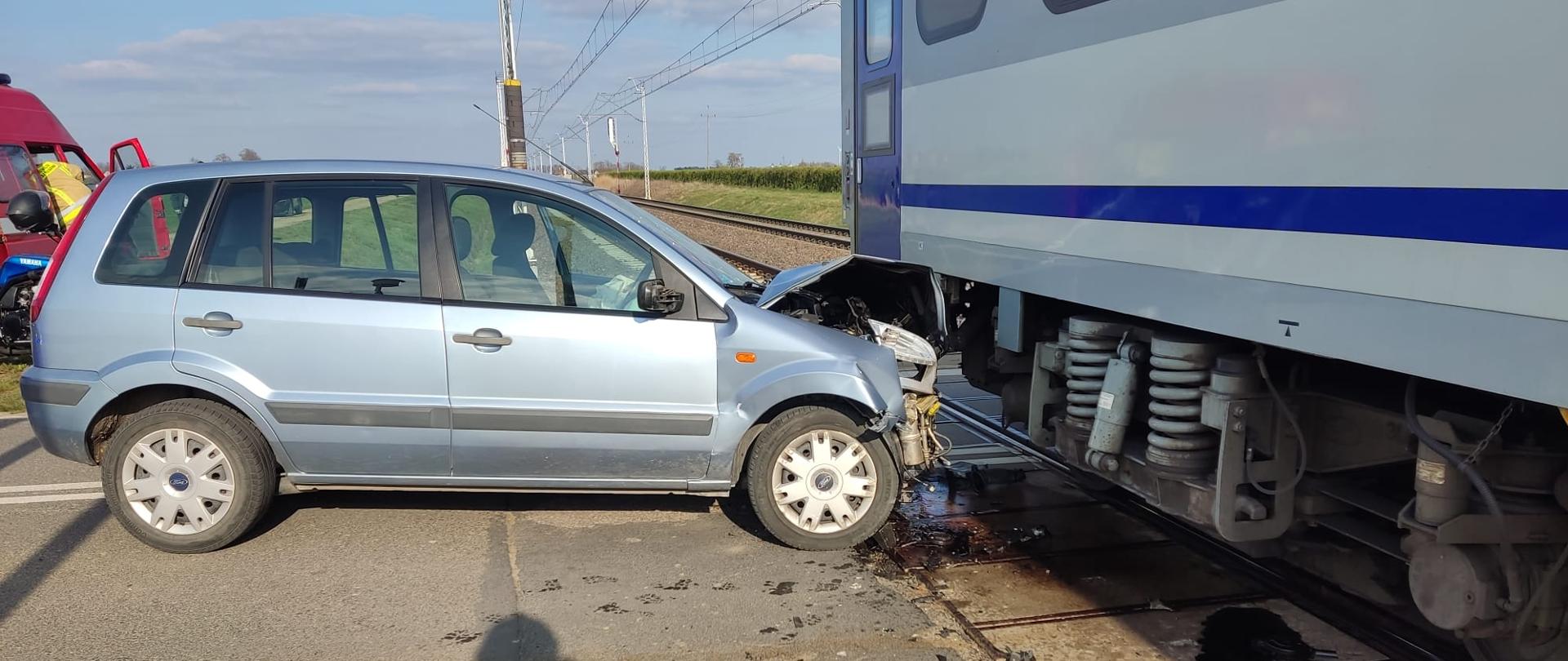Na zdjęciu widać miejsce wypadku na przejeździe kolejowym. Srebrny samochód typu hatchback, z poważnymi uszkodzeniami przedniej części, znajduje się tuż obok boku pociągu. Pociąg jest częściowo widoczny, widać jego szare i niebieskie wagony oraz podwozie z kołami stojące na torach. Samochód stoi na drodze przecinającej tory kolejowe, a jego przód jest zmiażdżony i wciśnięty pod pociąg. Wokół miejsca zdarzenia widać fragmenty uszkodzonych pojazdów i prawdopodobnie płyny eksploatacyjne na jezdni. Nad miejscem wypadku widać słupy z trakcją elektryczną. Niebo jest jasne, a dzień słoneczny. W oddali rozciąga się krajobraz z polami i zielenią.
