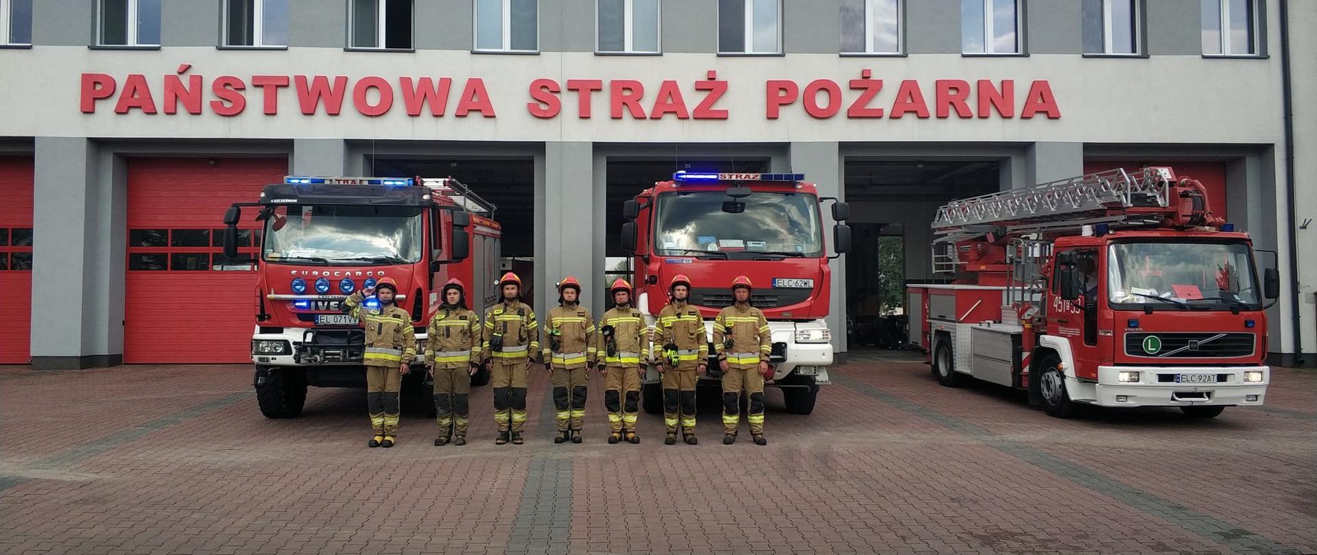 siedmiu strażaków ubranych w stroje specjalne stoi na tle trzech czerwonych samochodów ratowniczo gaśniczych za nimi znajduje się szary budynek z czerwonym napisem Państwowa Straż Pożarna
