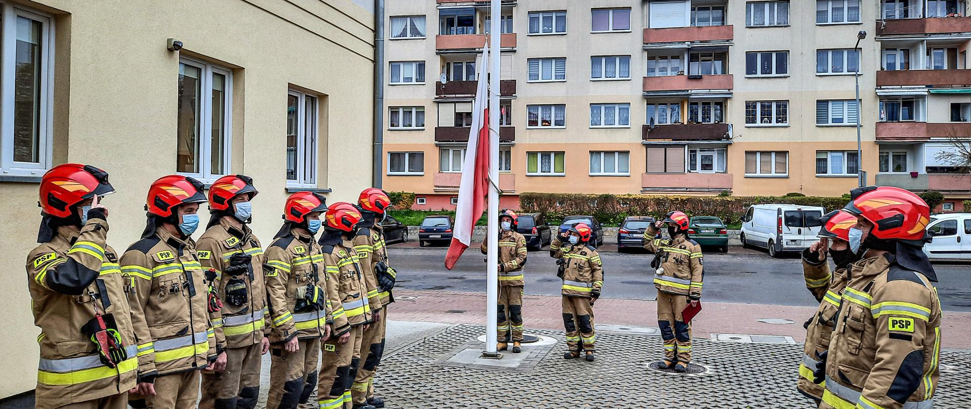 Na pierwszym planie zdjęcia znajdują się strażacy stojący w szeregu naprzeciwko siebie podczas zmiany służby. Dalej za nimi przy maszcie trzech strażaków zakłada i uroczyście podnosi flagę ma maszt. 