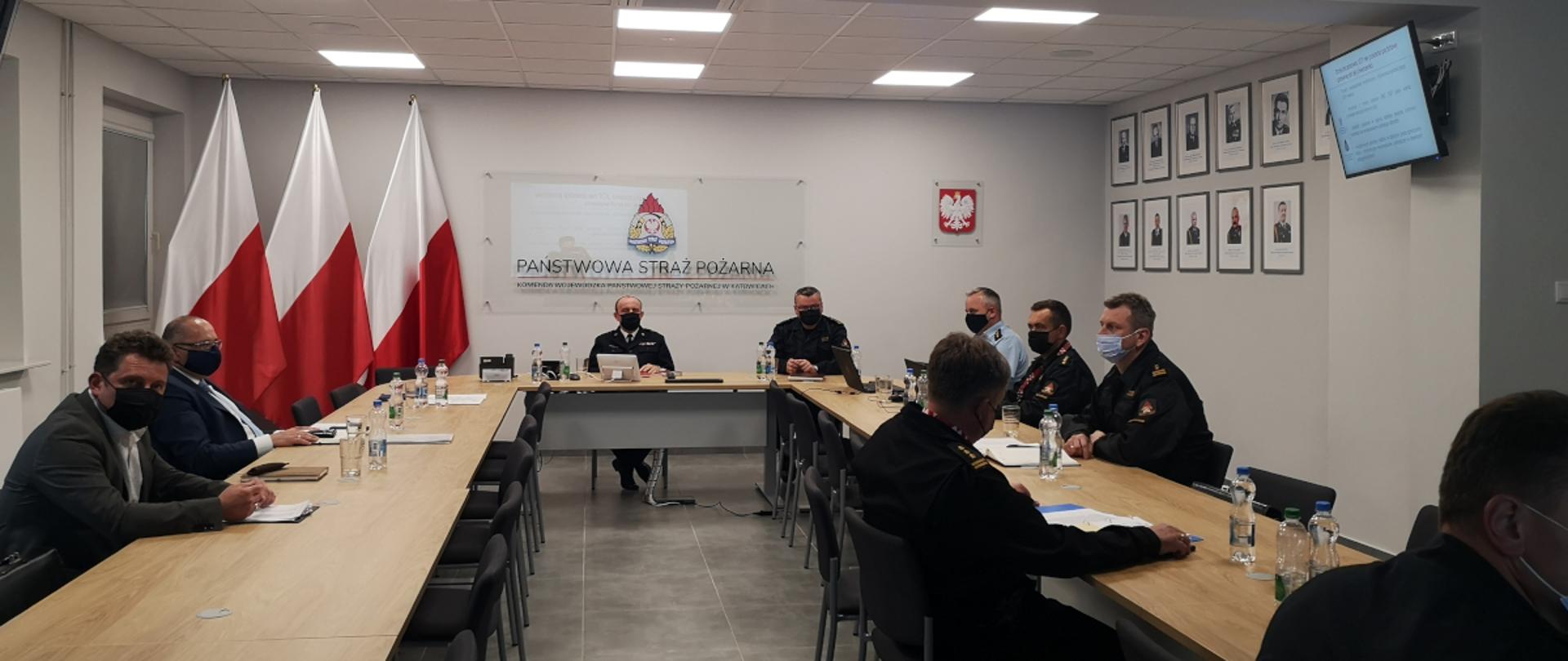 Przy stole ustawionym w kształcie litery U siedzą ubrani w maseczki ochronne przedstawiciele OPS, u szczytu stołu siedzą dwaj zastępcy Śląskiego Komendanta Wojewódzkiego PSP