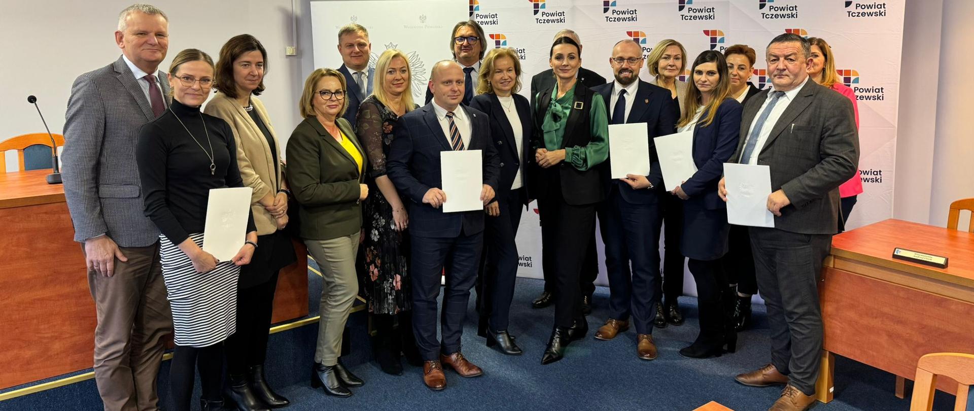 grupa osób stoi na tle baneru z napisem powiat tczewski, część trzyma w dłoniach teczki z podpisanymi umowami