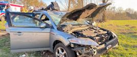 Na zdjęciu widoczny jest uszkodzony front pojazdu osobowego. W tle widoczna jest białą karetka pogotowia. Pojazd na poboczu drogi, na kołach. 