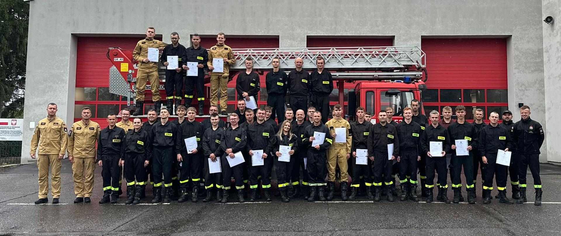 Grupa strażaków OSP w ubraniach koszarowych trzymająca zaświadczenia ukończenia kursu. Wśród strażaków OSP są również funkcjonariusze PSP. Wszyscy na tle samochodu pożarniczego typu podnośnik, przed budynkiem komendy powiatowej.
