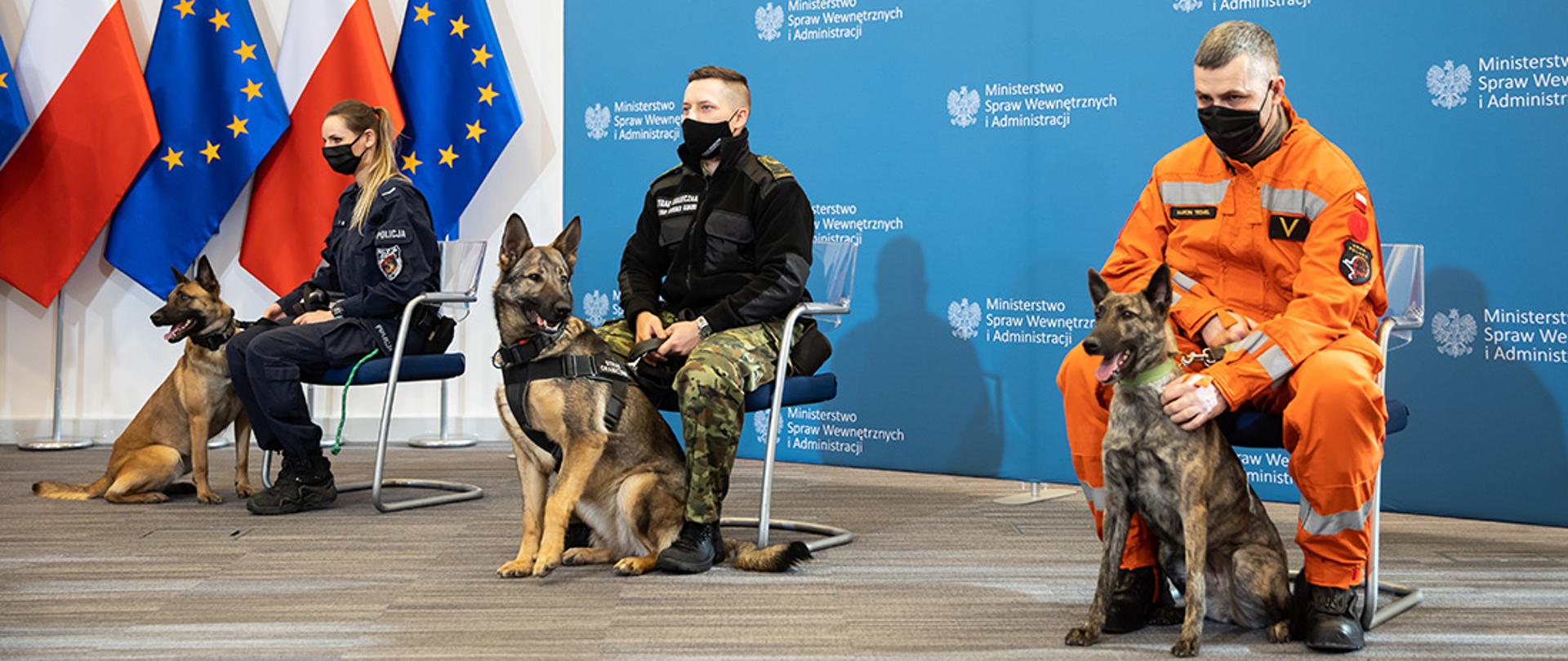 Zdjęcie w trakcie trwania konferencji prasowej w MSWiA. Na zdjęciu policjantka, strażnik graniczny i strażak - każdy u boku trzyma psa, który razem z nim służył w służbie. Funkcjonariusze są w mundurach. W pomieszczeniu flagi narodowe i baner MSWiA.