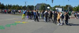 Ćwiczenia ewakuacyjne. Boisko Szkoły Podstawowej w Sierakowie. Uczniowie pod nadzorem nauczycieli opuścili budynek szkoły i udają się w wyznaczone miejsce zbiórki, gdzie ustawiają się klasami. W tle drzewa, lampy i budynki.