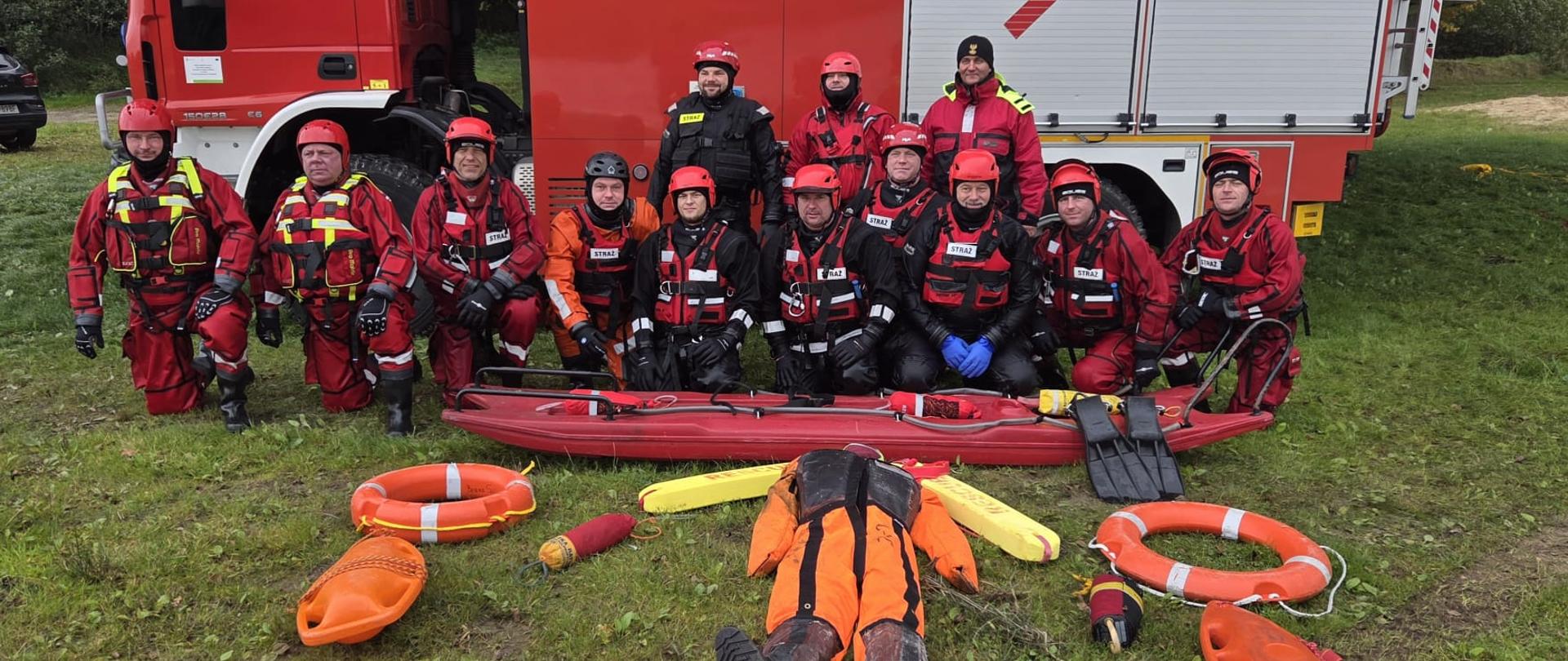 Grupa strażaków w ubraniach do działań w wodzie pozuje przed samochodem ratowniczym typu ciężkiego. Na trawie leżą manekiny, koła ratunkowe i sprzęt do ćwiczeń z ratownictwa wodnego.