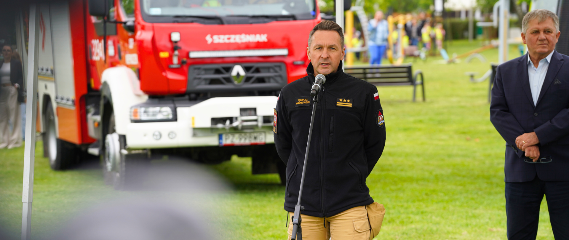 Przedstawiciele służb stoją pod namiotem na trawie, przygotowując się do briefingu prasowego. W tle samochód straży pożarnej - głos zabrał strażak, stojąc przy mikrofonie.