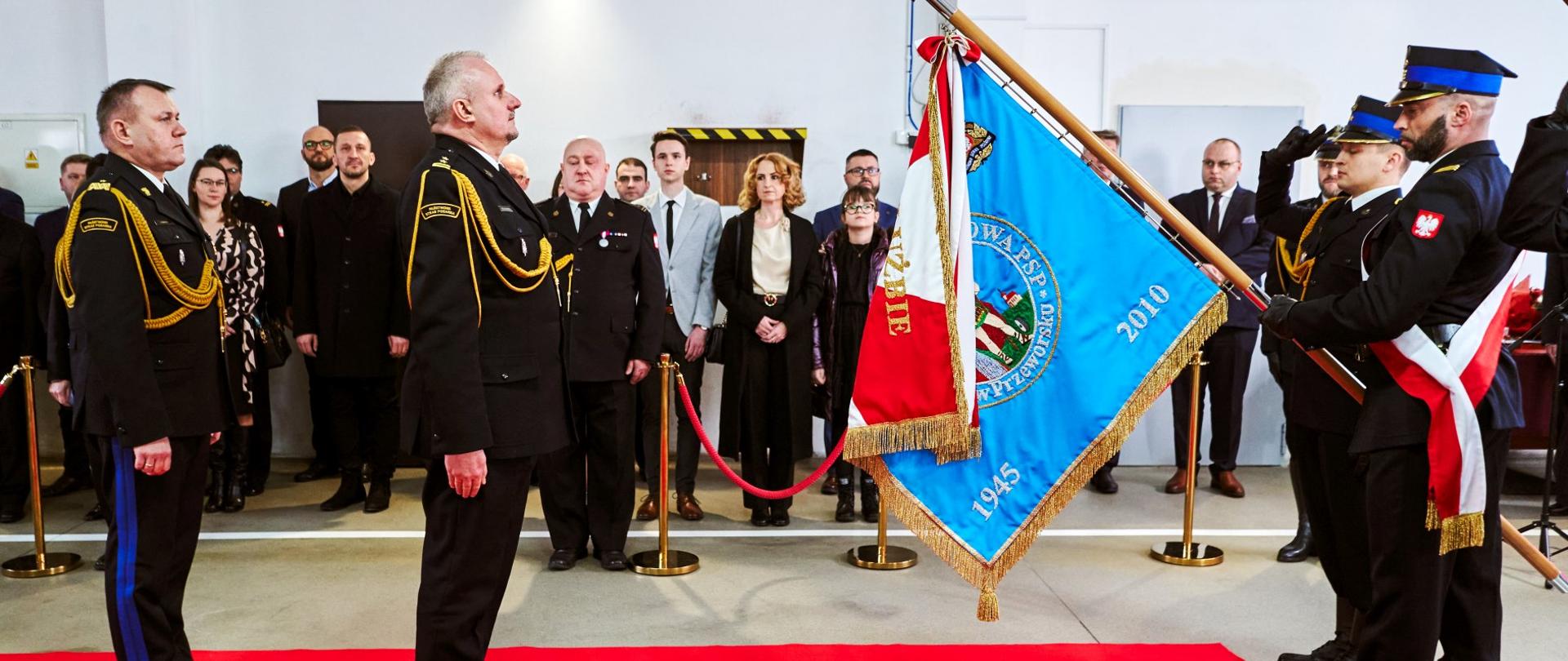 Zdjęcie zrobione w pomieszczeniu, w garażu jednostki ratowniczo-gaśniczej, podczas uroczystego apelu. Poczet sztandarowy trzyma pochylony sztandar. Przed sztandarem stoją oficerowie Państwowej Straży Pożarnej. Z tyłu stoją zaproszeni na uroczystość goście.