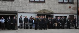 Grupa strażaków w galowych mundurach oraz zaproszonych gości stoi przed budynkiem podczas uroczystości strażackiej. W tle widoczne są krzesła przygotowane dla uczestników wydarzenia oraz fragment budynku remizy.