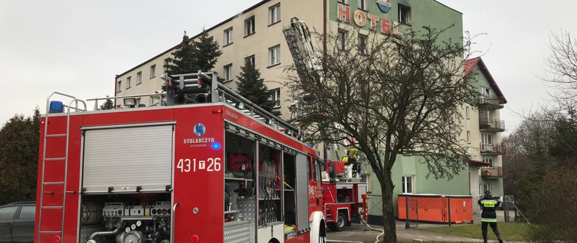 Tył samochodu gaśniczego z rozwiniętymi liniami gaśniczymi w stronę hotelu. W tle za nim podnośnik hydrauliczny i hotel od strony południowo-zachodniej.