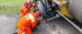 Zdjęcie przedstawia dwóch strażaków grupy poszukiwawczej (SGPR "Brzeg") w kombinezonach koloru pomarańczowego z czerwonymi kaskami na głowach. Strażacy w pozycji klęczącej przy pomocy kamery wziernikowej sprawdzają przestrzenie puste pod przewróconą cysterną leżącą po prawej stronie kadru. Strażacy klękają na asfalcie, w dalszym planie trawa. Po lewej stronie widoczny kawałek drabiny przenośnej przystawnej.