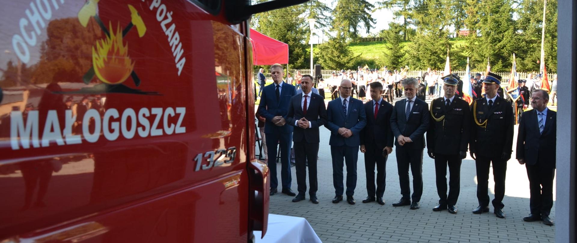 Zdjęcie po lewej stronie przedstawia przednią część samochodu ratowniczo-gaśniczego z napisem na drzwiach Ochotnicza Straż Pożarna Małogoszcz. Na drugim planie widać ustawionych obok siebie zaproszonych gości, którzy przyglądają się nowo otwartej strażnicy.