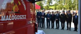 Zdjęcie po lewej stronie przedstawia przednią część samochodu ratowniczo-gaśniczego z napisem na drzwiach Ochotnicza Straż Pożarna Małogoszcz. Na drugim planie widać ustawionych obok siebie zaproszonych gości, którzy przyglądają się nowo otwartej strażnicy.