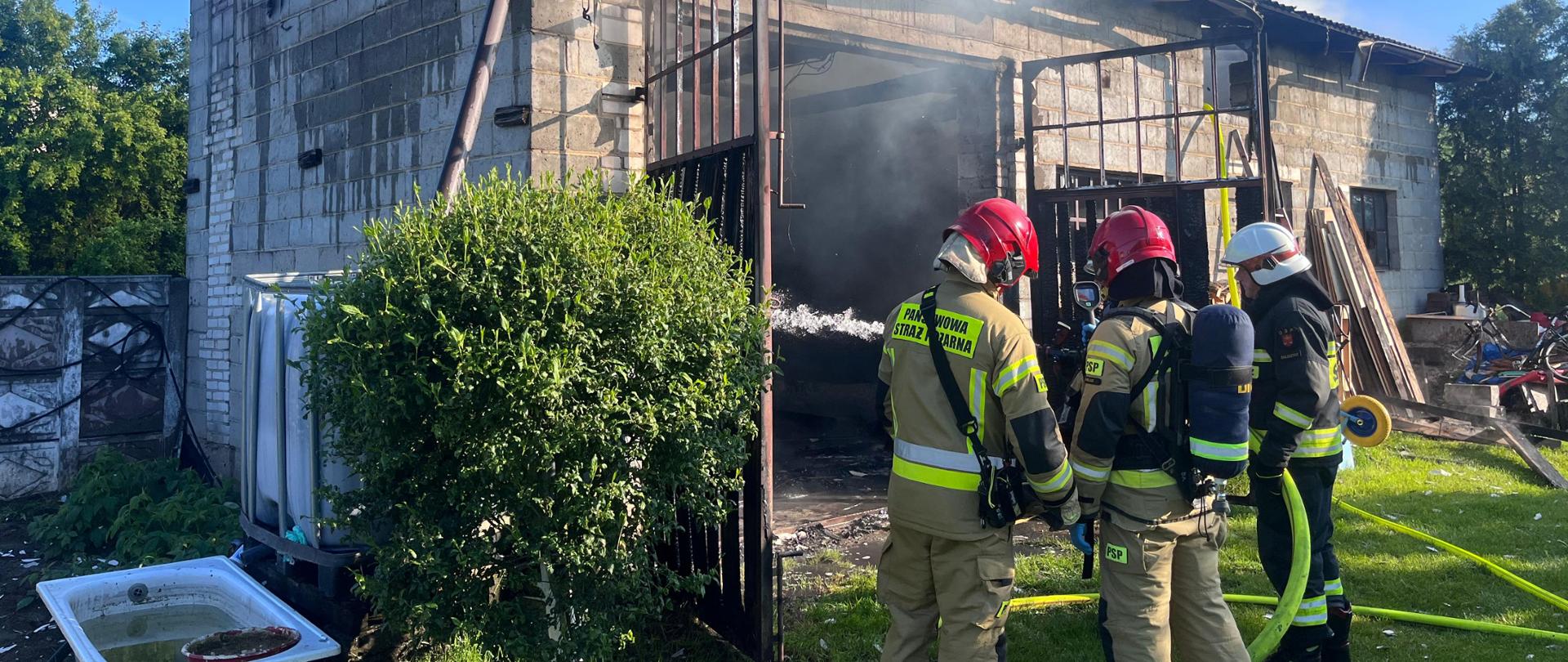Zdjęcie przedstawia budynek gospodarczy gdzie doszło do pożaru. Przed otwarta bramą stoi trzech strażaków.