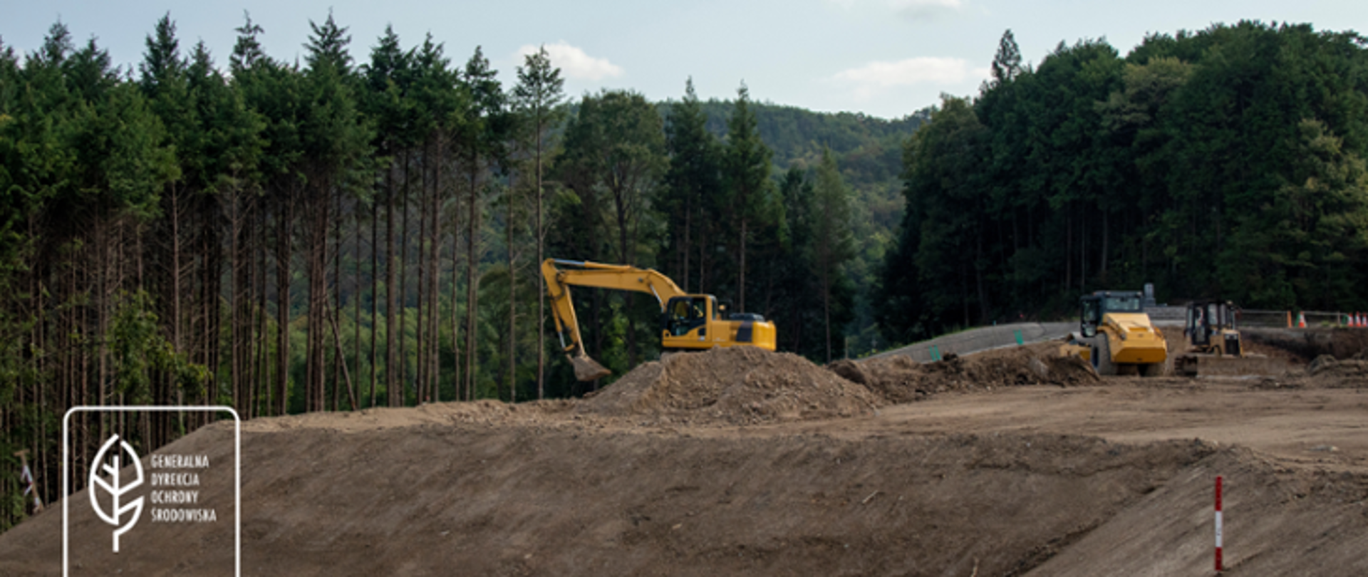 W tle las. Dwie maszyny budowlane (koparki) pracują na nasypie ziemnym. W lewym dolnym rogu logo generalnej Dyrekcji Ochrony Środowiska (biały liść).