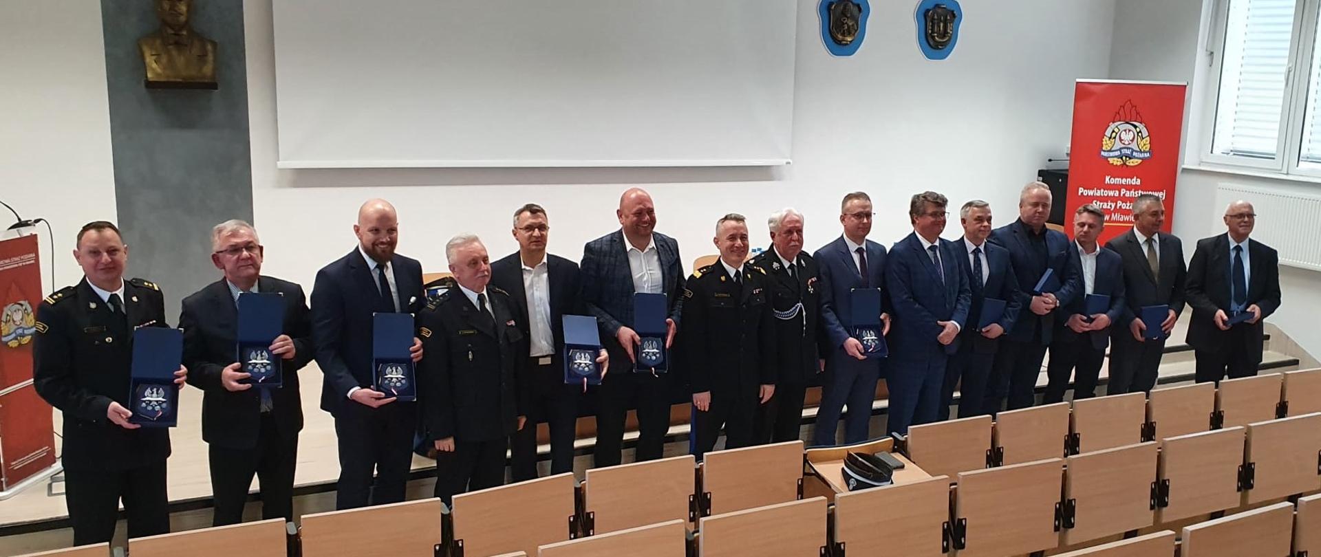 Funkcjonariusze i osoby cywilne stoją w rzędzie w auli podczas odprawy rocznej