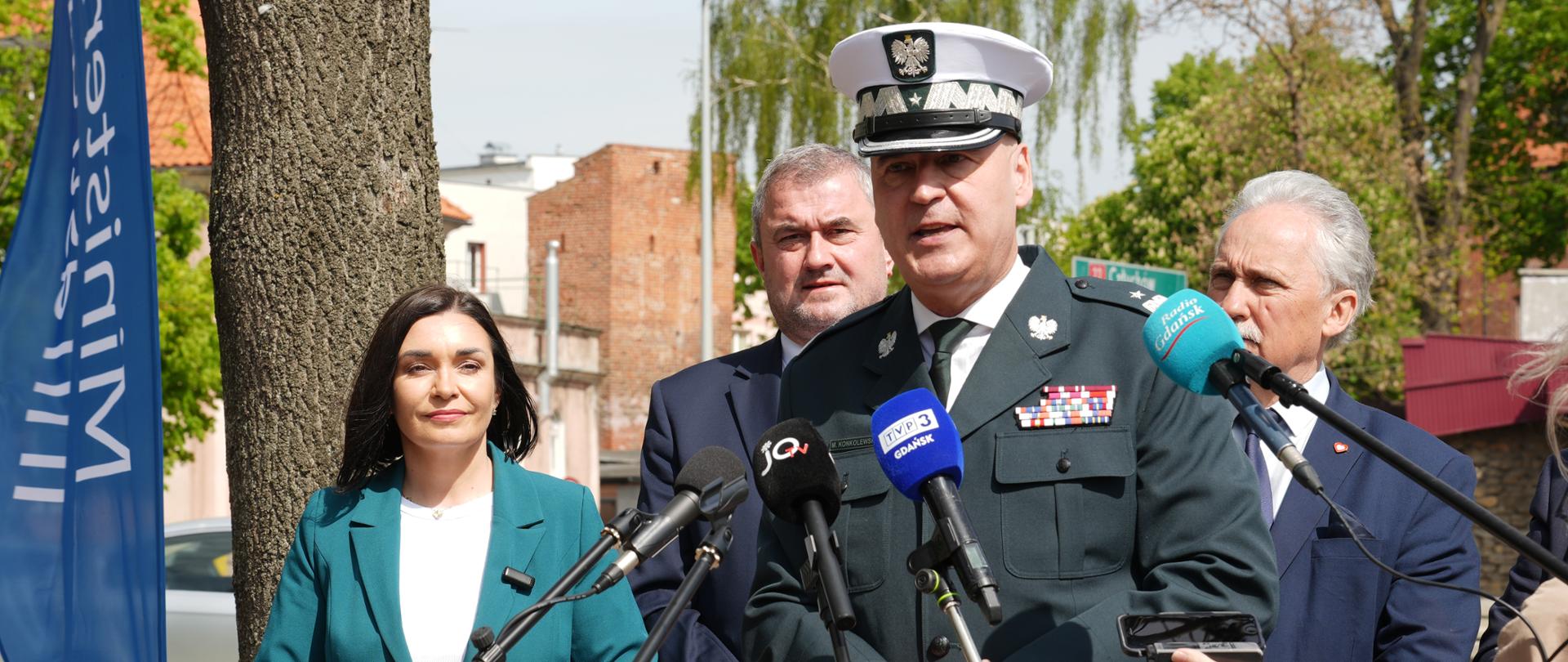 Mężczyzna w mundurze przemawia do mikrofonu. Za nim stoją: kobieta w niebieskiej marynarce i dwaj mężczyźni w garniturach.