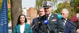 Mężczyzna w mundurze przemawia do mikrofonu. Za nim stoją: kobieta w niebieskiej marynarce i dwaj mężczyźni w garniturach.