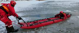 Na zdjęciu widać dwóch strażaków-ratowników na lodzie. Obaj ubrani są w czerwone skafandry i kaski. Jeden strażak trzyma się czerwonych plastikowych sań, drugi trzyma sanie stojąc na lodzie. W oddali widać zamarznięty akwen wodny i zalesiony brzeg.
