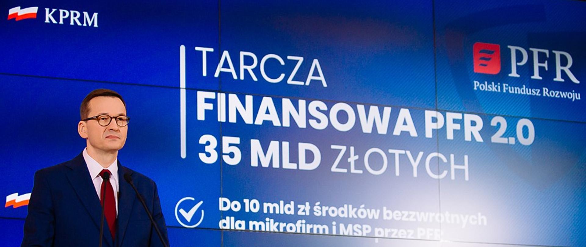 Premier Mateusz Morawiecki stoi przy mównicy, w tle prezentacja na ekranie.
