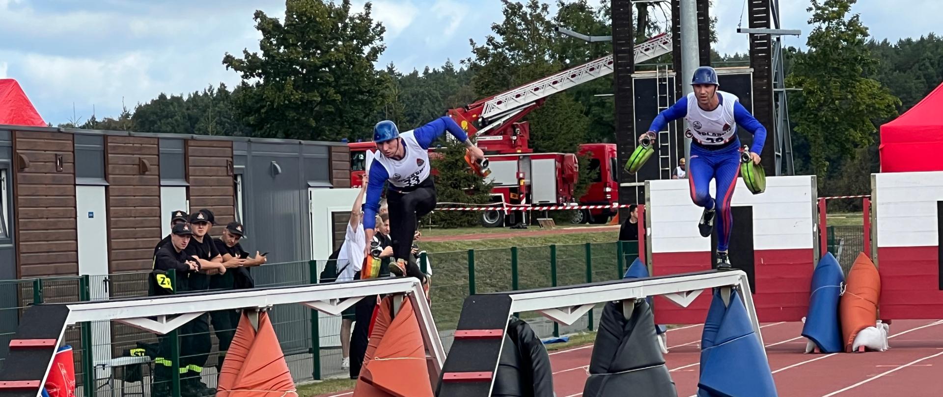 Dwóch strażaków ubranych na sportowo biegnie po torze po dwóch równoważniach. W rękach trzymają węże. 