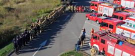 Na zdjęciu widoczni strażacy oraz samochody ratowniczo-gaśnicze podczas odprawy przed rozpoczęciem ćwiczeń. Strażacy stoją w dwuszeregu.