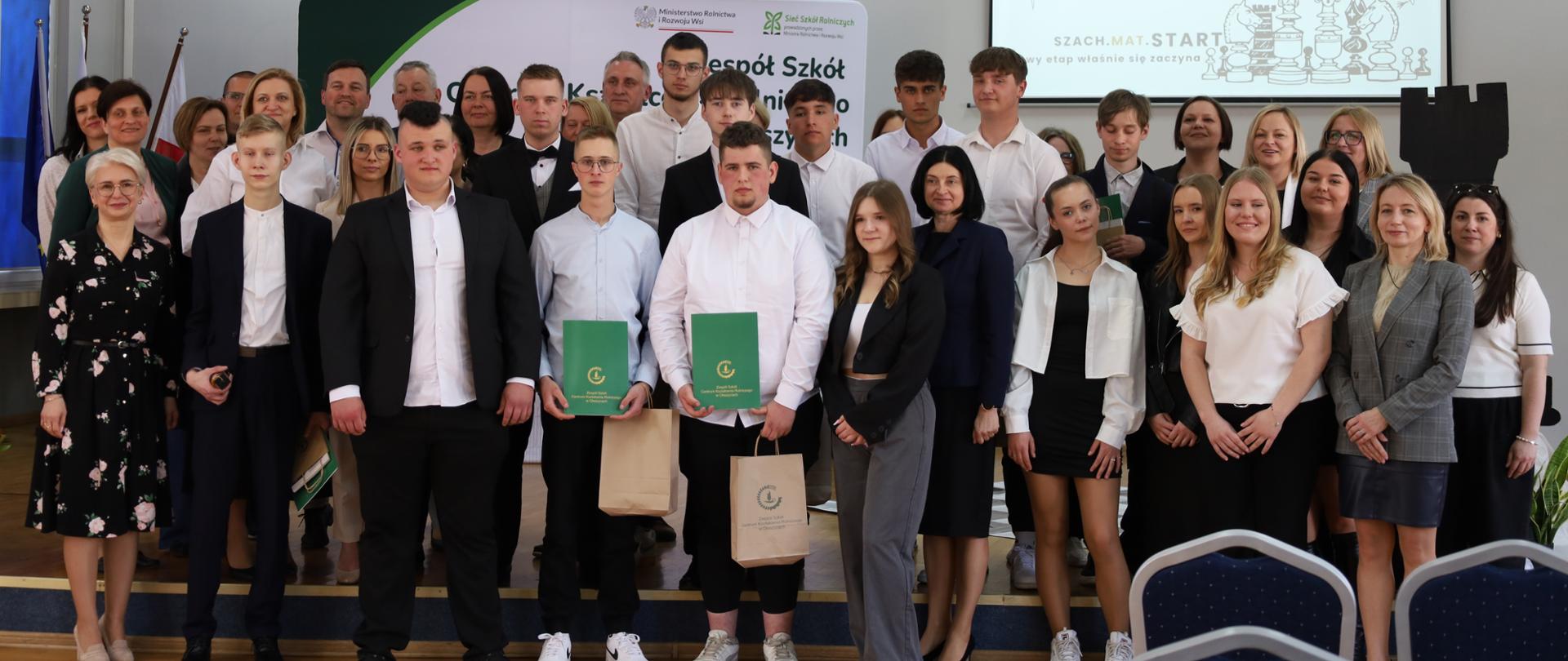 Zdjęcie grupowe. Na scenie stoją uczniowie klasy maturalnej i nauczycieli. 