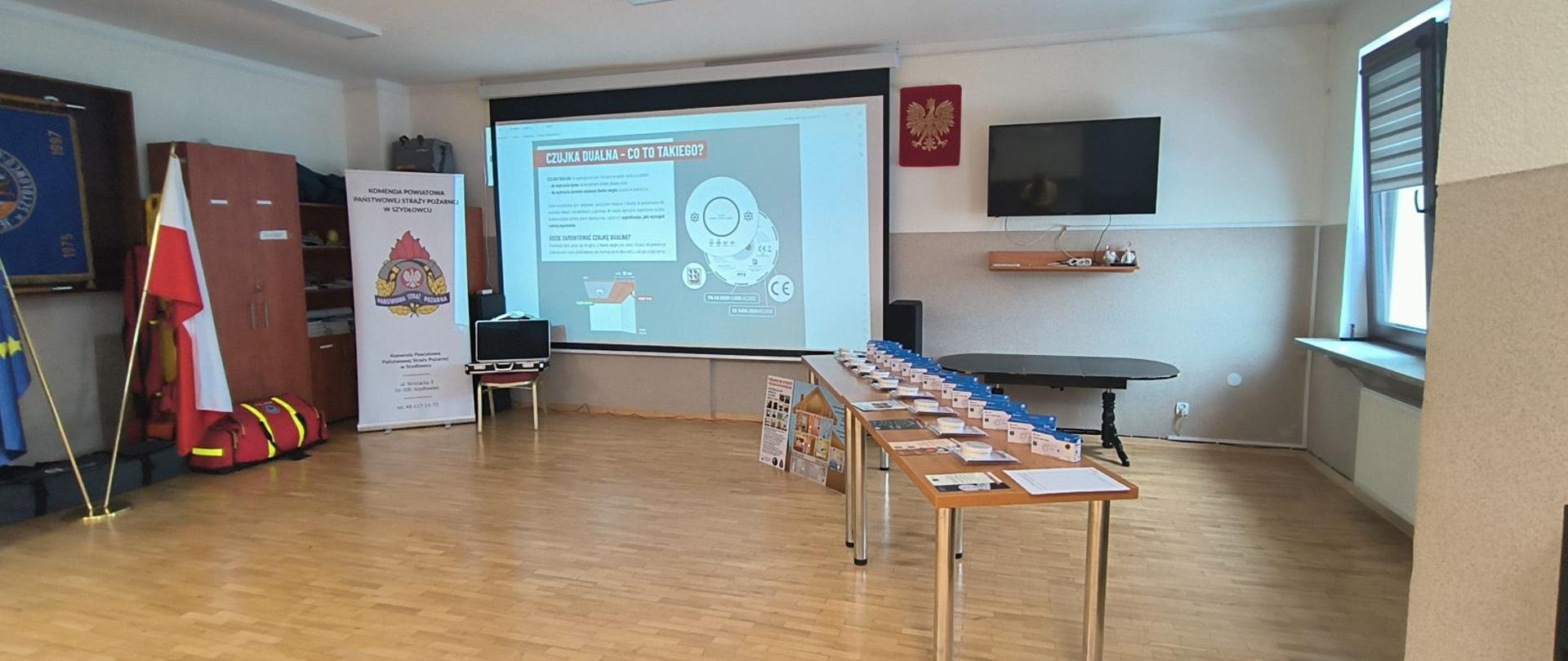 Zdjęcie przedstawia wnętrze sali konferencyjnej przygotowanej do kampanii edukacyjnej z zakresu bezpieczeństwa przeciwpożarowego. Pomieszczenie jest jasne, z drewnianą podłogą, utrzymane w oficjalnym, instytucjonalnym stylu.