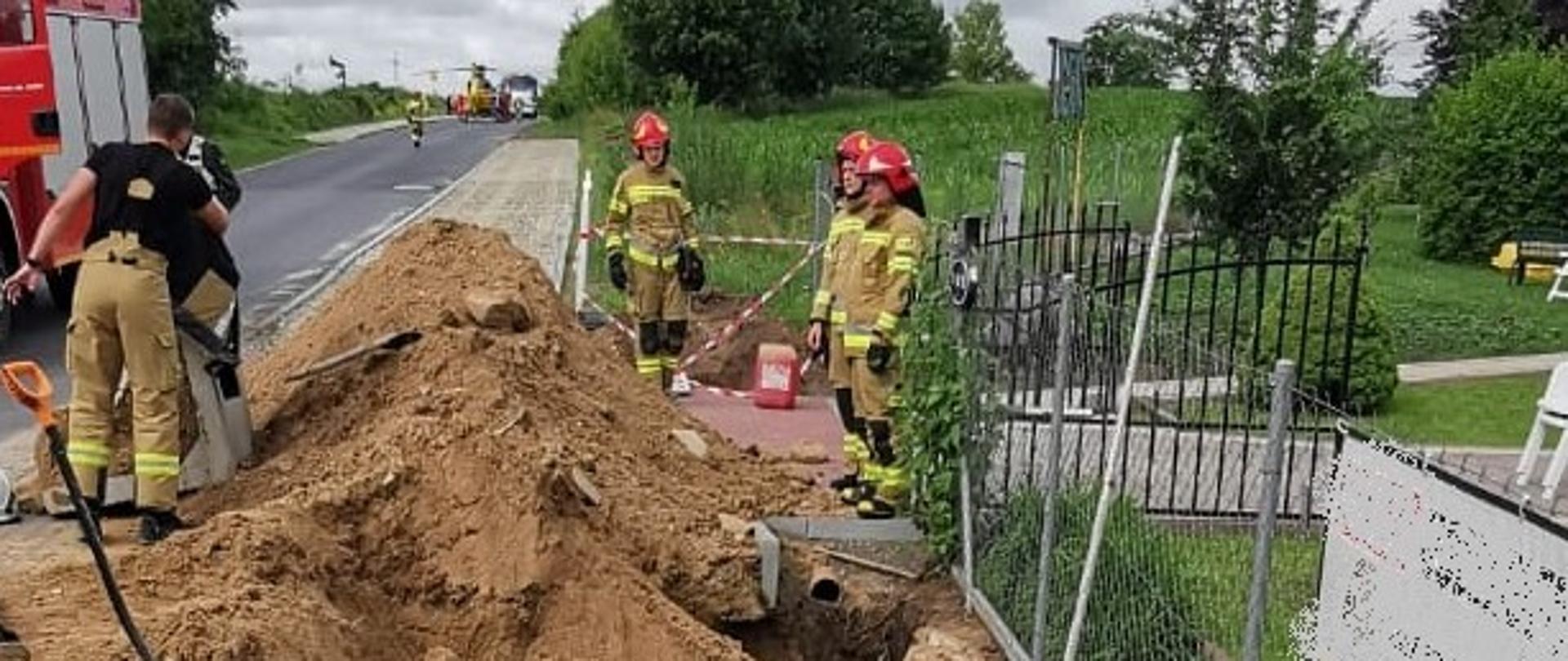 Zdjęcie przedstawia wykop pod kanalizację deszczową przy drodze. Na zdjęciu widać ziemię, chodnik i drogę oraz strażaków i samochód strażacki. 