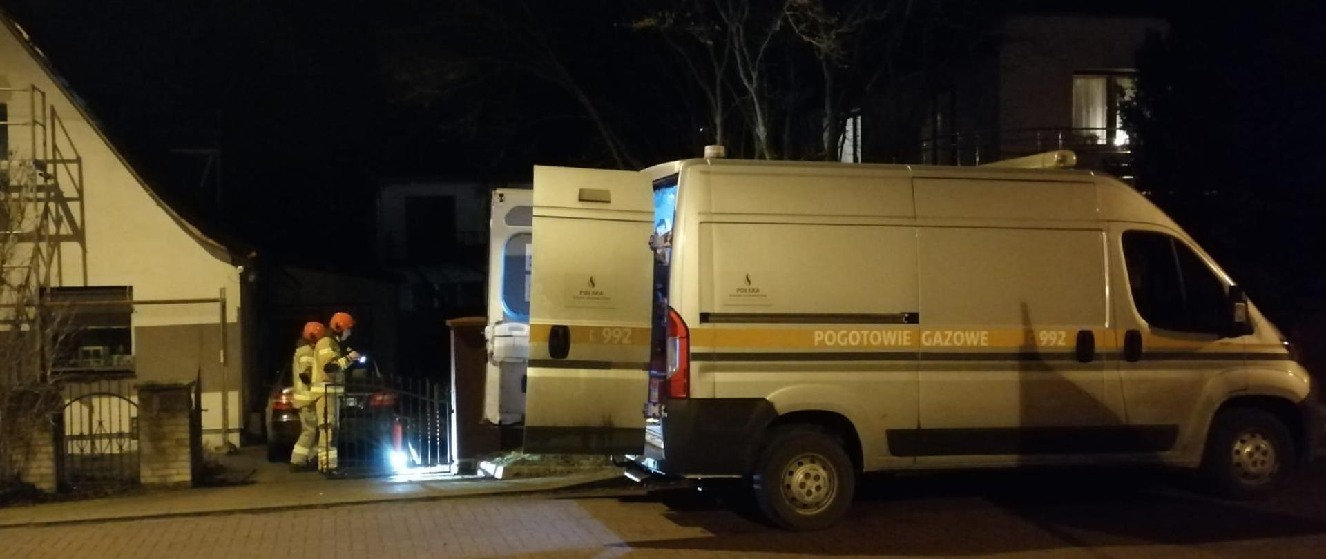 Zdjęcie przedstawia drogę z kostki brukowej na której stoi biały bus z napisem Pogotowie Gazowe. Po lewej stronie stoją strażacy w piaskowych ubraniach specjalnych. Na trzecim planie znajduje się dom jednorodzinnym przy którym rozstawione jest rusztowanie. Po podwórku chodzą i świecą latarkami. Zdjęcie zrobiono porą nocną.