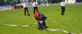 Zdjęcie przedstawia druha, który podczas zawodów OSP łączy ze sobą dwa odcinki węża. Druh na kolanach. Z tyłu widać sędziów. 