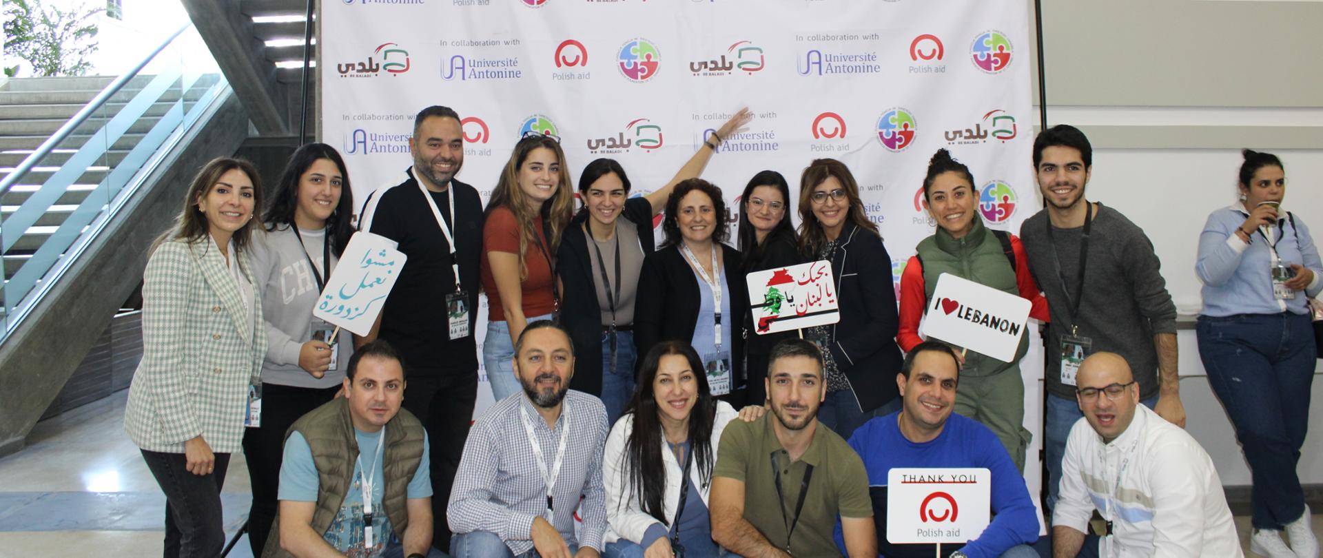 Grupa kobiet i mężczyzn w różnym wieku pozuje na tle roll-upa z logo Polska pomoc, Universite Antonine i Be Baldi