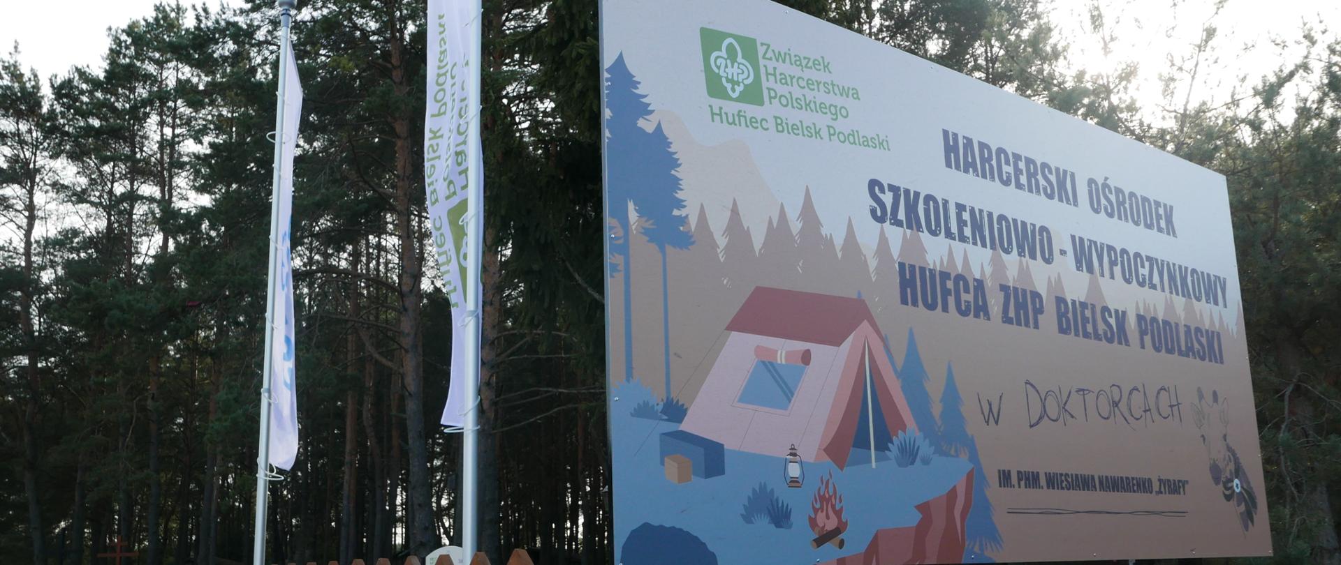 Kolorowa tablica informacyjna Hufca ZHP Bielsk Podlaski w Doktorcach. W lewym górnym rogu widzimy harcerską krajkę, pod spodem namiot harcerski, przed nim ognisko i lampa naftowa. Na płocie białe i błękitne baloniki. Za płotem las.