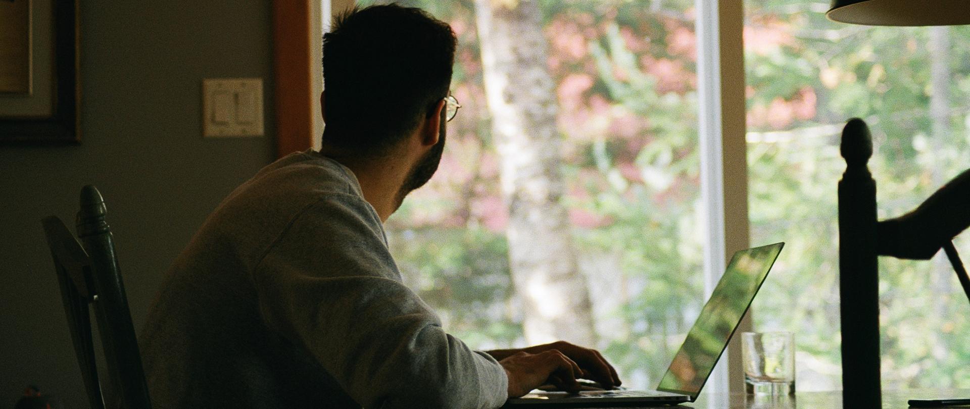 Mężczyzna siedzi w domu przy stole z otwartym laptopem. Wygląda przez okno
