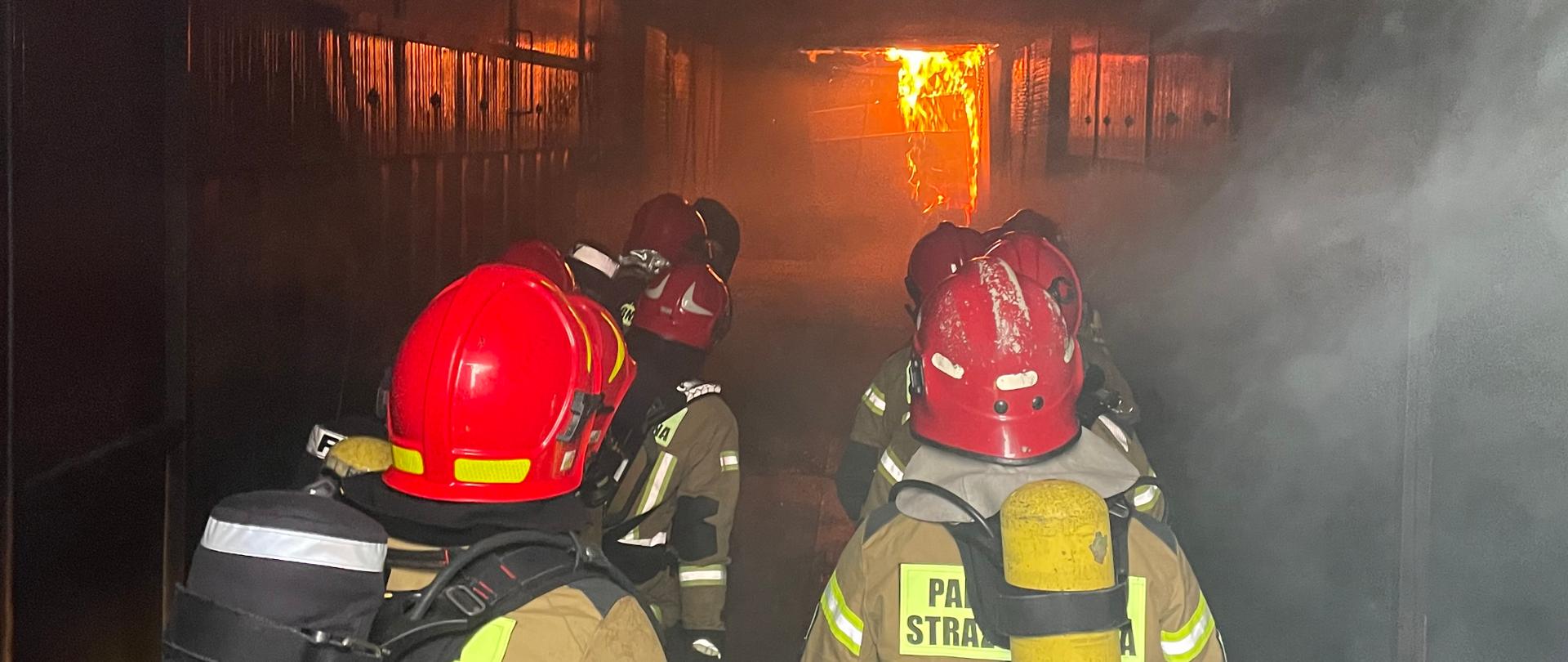 Na zdjęciu widać znajdujących się w pomieszczeniu kilku klęczących strażaków odwróconych tyłem. Ludzie obserwują pożar znajdujący się przed nimi. Mają na sobie aparaty ochronne układu oddechowego, czerwone hełmy oraz ubrania ochronne w kolorze piaskowym. Nad nimi unosi się dym. 