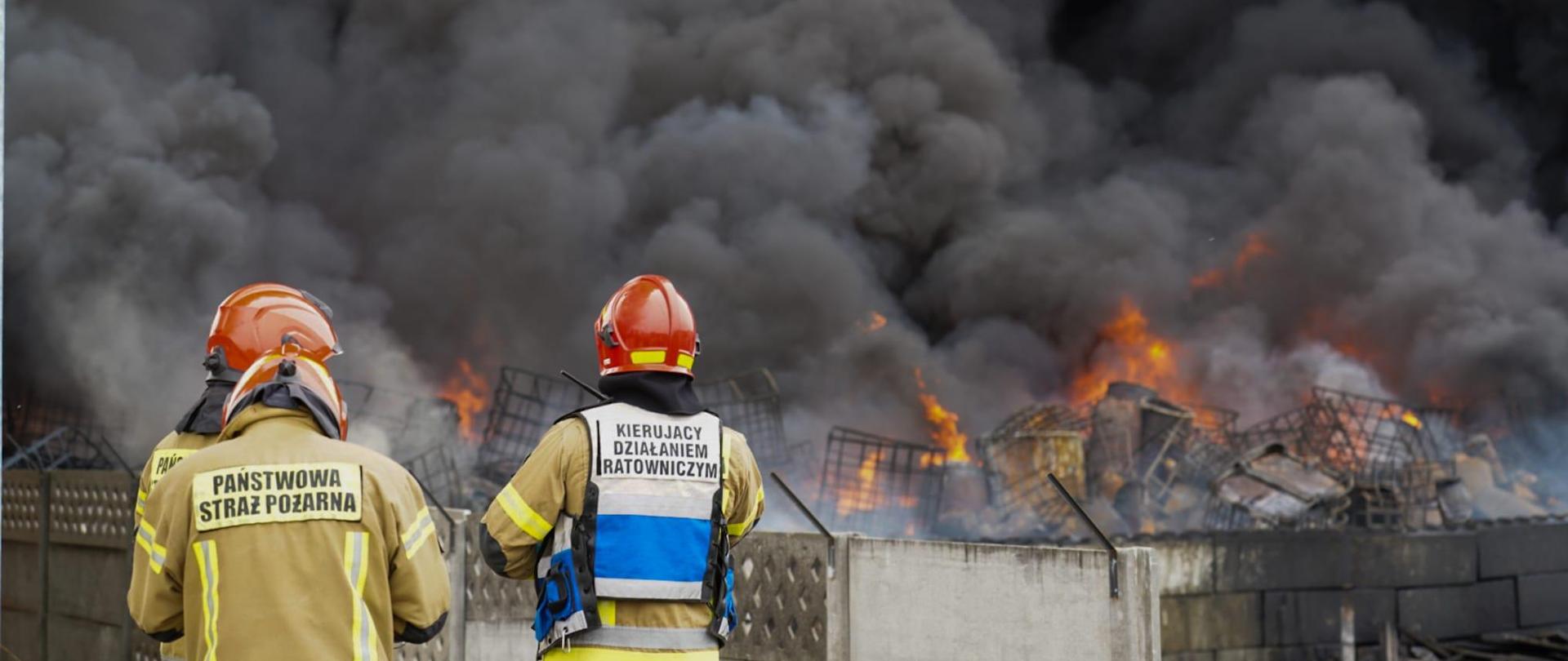 Trzech strażaków w tym kierujący działaniem ratowniczym nadzorują działania straży pożarnej podczas pożaru. 