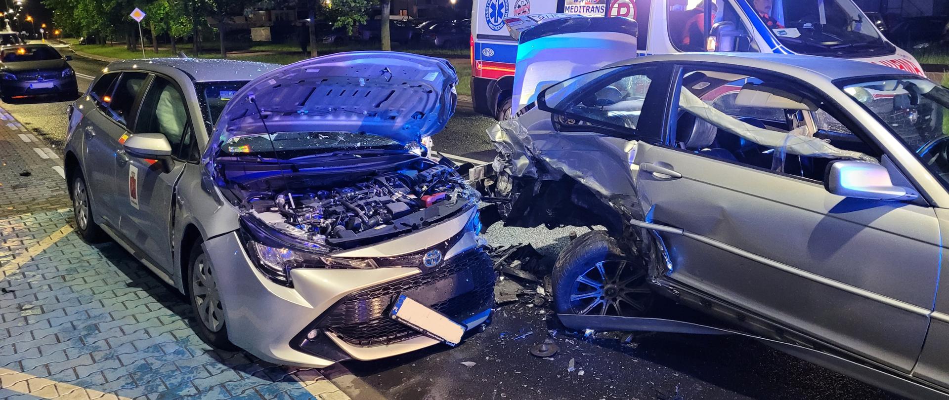 Na zdjęciu widoczne są dwa auta po wypadku. Widać znaczne zniszczenia pojazdów. W tle widoczna jest karetka z włączonymi sygnałami świetlnymi. Za karetką widać drzewa a na nich liście.
