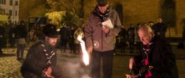 Kapelan harcerzy ksiądz Józef Nowakowski, Prezydent Miasta Torunia Pan Michał Zaleski i Komendant Hufca ZHP Toruń phm. Adam Wiznerowicz rozpalają ognisko światłem światłem Betlejemskim na Rynku Nowomiejskim w Toruniu w tle widać budynek Tumultu.
