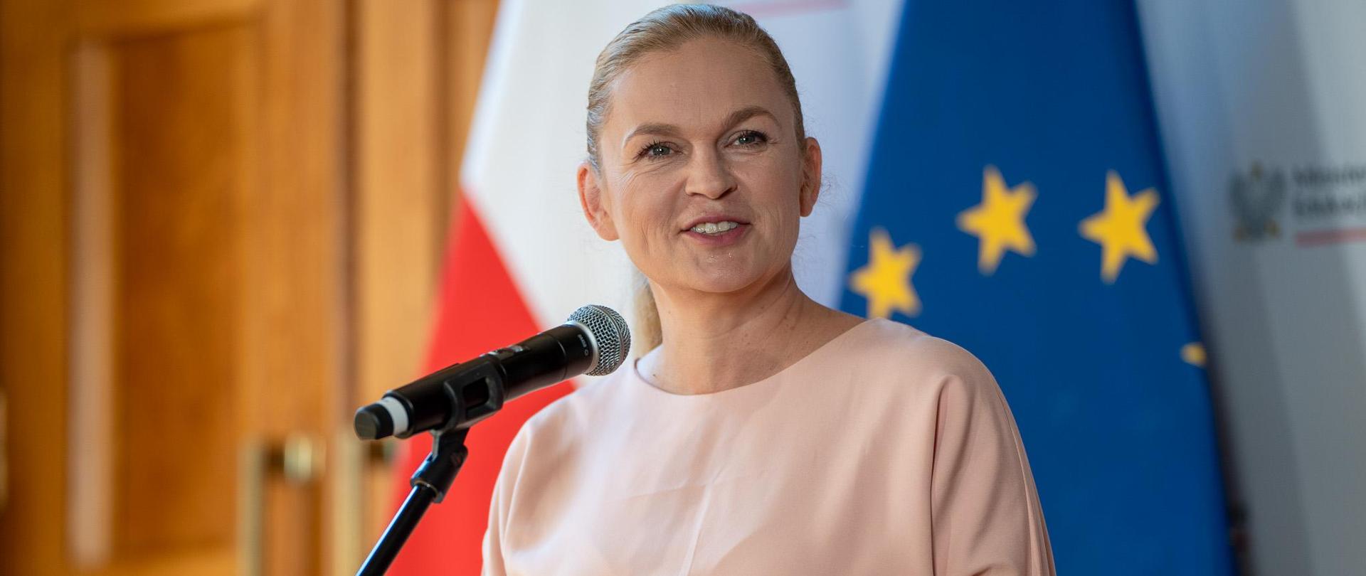 Minister Barbara Nowacka stoi przy mikrofonie i mówi, w tle flagi Polski UE