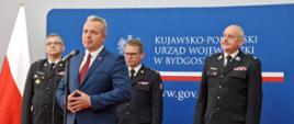 Na pierwszym planie wojewoda kujawsko-pomorski, za nim strażacy w mundurach wyjściowych - dwóch PSP, jeden OSP. Za plecami ścianka medialna z logo Urzędu Wojewódzkiego.