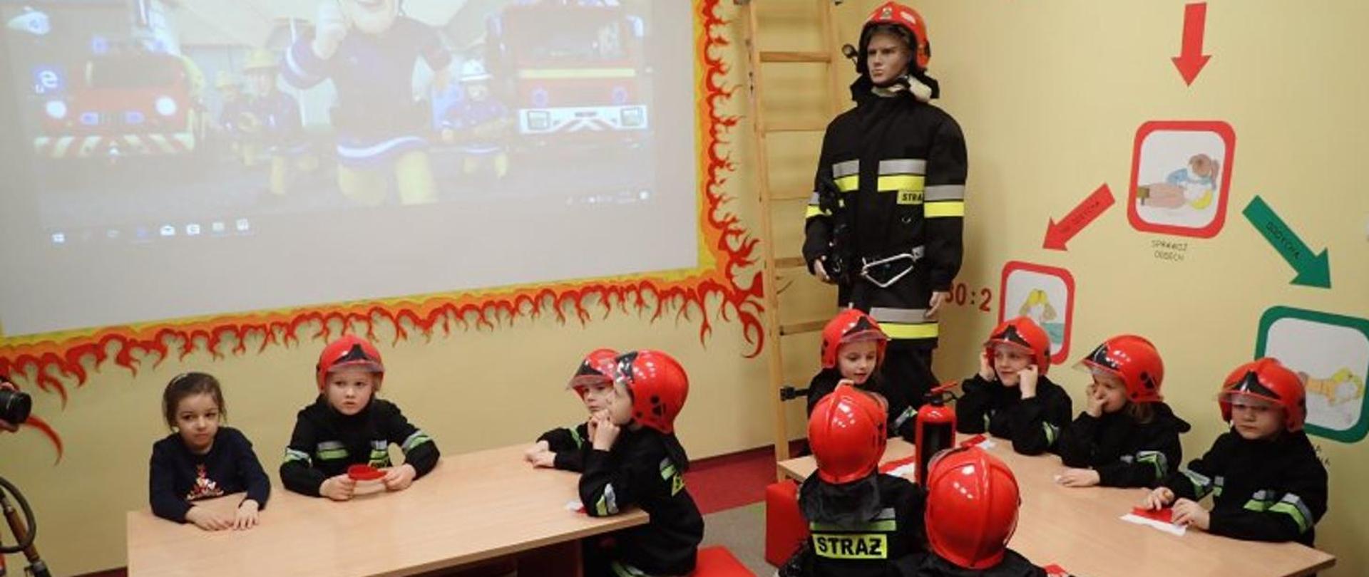 Zdjęcie przedstawia grupę dzieci w czerwonych hełmach strażackich, które siedzą w sali lekcyjnej. W centralnej części sali stoi manekin strażaka w umundurowaniu specjalnym, a obok znajduje się fragment ekranu, na którym wyświetlana jest kreskówka z motywem strażackim.