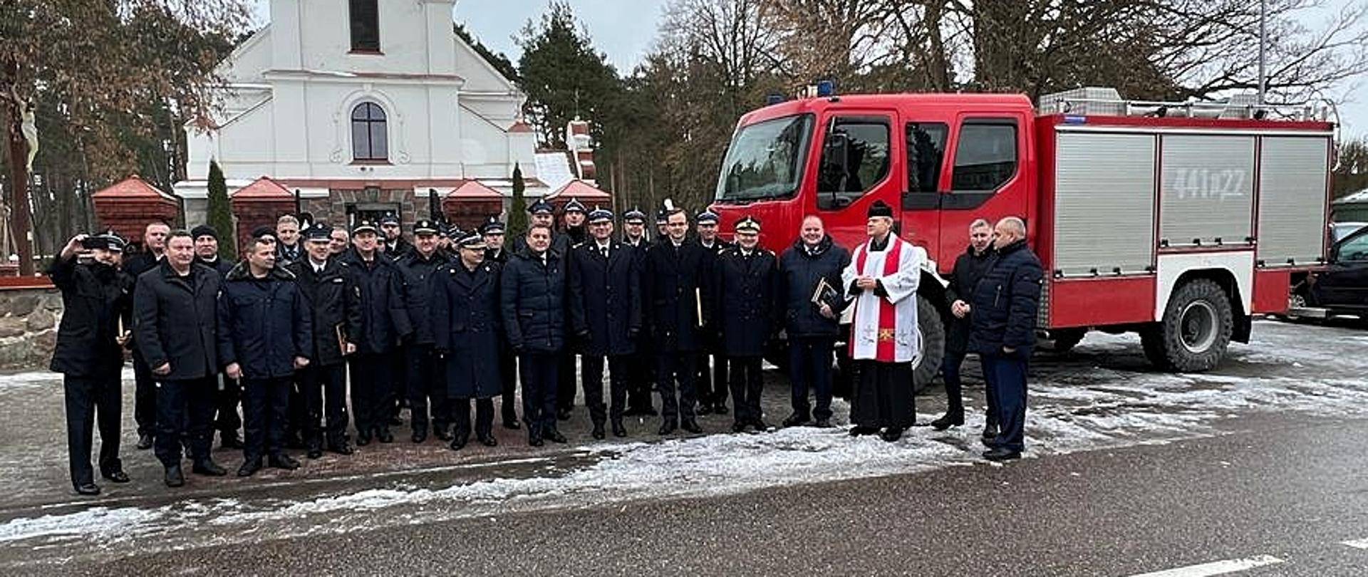 Zdjęcie grupowe uczestników uroczystości przy przekazywanym samochodzie. W tle budynek kościoła