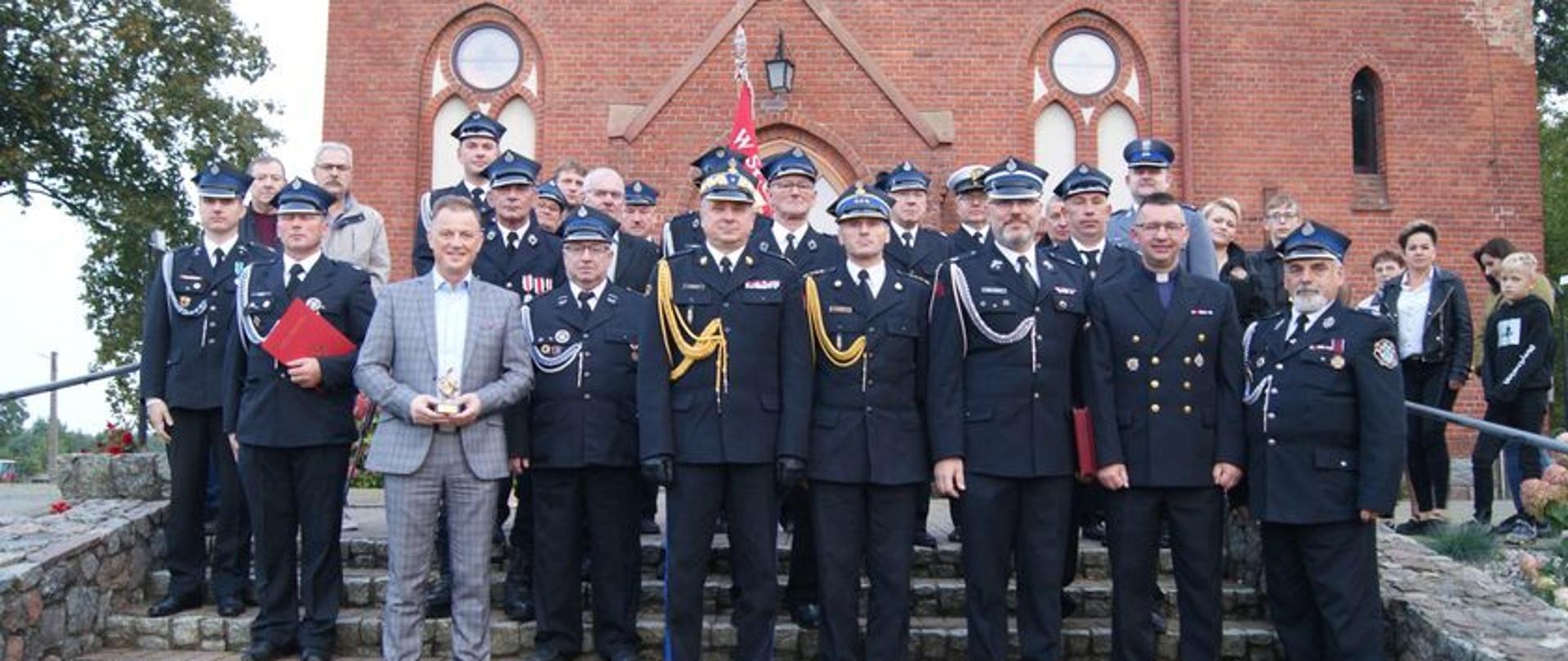 Na zdjęciu widać strażaków ubranych w mundury wyjściowe na tle kościoła w Leśniewie